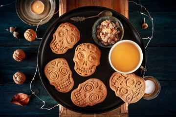 Halloween ginger cookies in the shape of skulls, homemade Dia de los muertos biscuits, with tea and candles, shot from above on a dark wooden background
