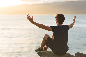 Uplifted young man feeling good sitting on top of a cliff above the ocean view and raising arms up...