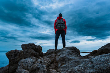 A man in a red jacket with a backpack on his back stands on the sea rocks and looks at the sea...