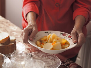 An elderly woman in brightly colored clothes sets the table, holds, and offers homemade diet potato soup as she treats guests.Healthy natural food.