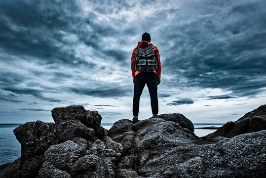 A man in a red jacket with a backpack on his back stands on the sea rocks and looks at the sea against the backdrop of storm clouds