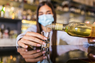 Beautiful female bartender with protective face mask pouring alcohol into shot glass during coronavirus pandemic, shelves full of bottles with alcohol on the background