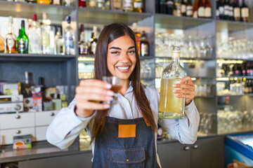 Beautiful female bartender is holding a shot glass with alcohol drink and a bottle in other hand, looking at camera and smiling while standing near the bar counter in cafe