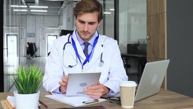 Doctor in white medical uniform with statoscope reading personal file of patient on tablet and writes data to laptop.