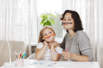 Obraz na płótnie Canvas Mother and daughter draw together. A charming girl and her cheerful mother play with colored markers while completing a task. Home training, online training