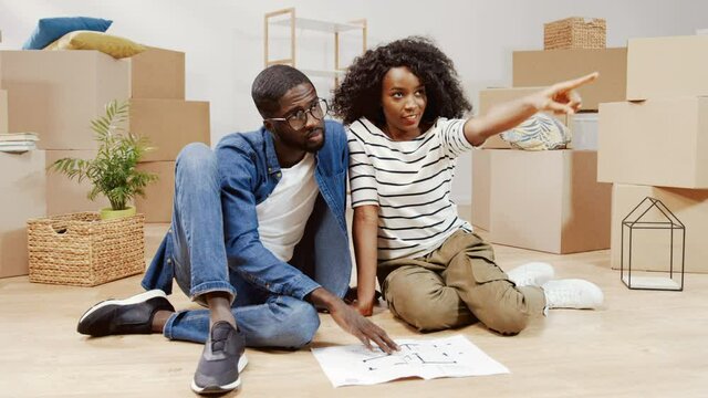 Portrait of young couple in new apartment. African-American man and woman are sitting on the floor and looking at layout of apartment and planning arrangement of furniture.