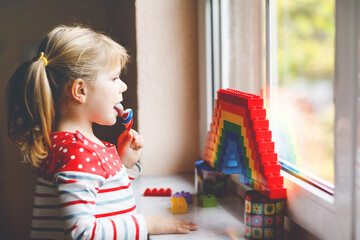 Cute little toddler girl by window create rainbow with colorful plastic blocks during pandemic coronavirus quarantine. Children made and paint rainbows around the world as sign. Kid eating lollipop.