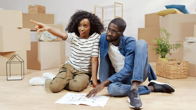 Portrait of young couple in new apartment. African American man and woman sitting on the floor and watching plan of apartment agree on arrangement of furniture.