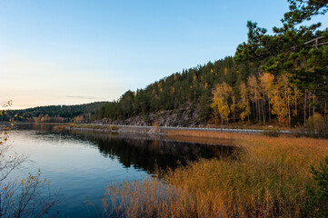Fototapeta na wymiar It's a beautiful sunset scenery. A lake with rocky shores and autumn forest on hilly terrain.