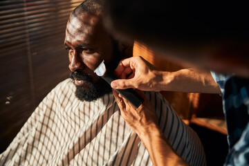 Barber shaving client beard with electric trimmer in barbershop