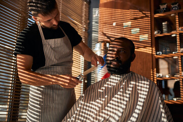 Male barber in apron trimming client beard in barbershop