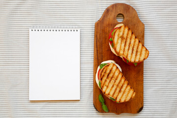 Tasty Grilled Caprese Panini (Mozzarella, Tomatoes and Basil) on a rustic wooden board, blank notepad, top view. Flat lay, overhead, from above. Copy space.