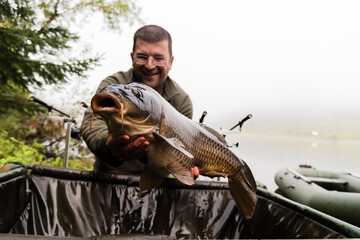 Happy smiling fisherman holding his fish  with lake on the background. Selective focus on the carp.