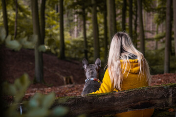 Young blonde woman in yellow coat and her dog sitting on a bench, looking into the autumn forest