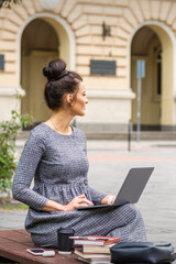 Student young woman uses laptop on the bench on the city street near university