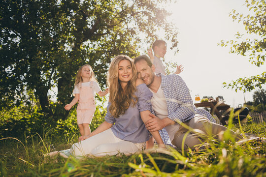 Full body size photo of full big family four people lovers mom dad two small kids beaming shiny smile cuddle sit carpet enjoy beautiful sunny day green garden home park backyard outside