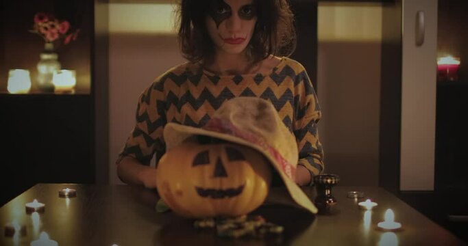 Young female with scary make up mask sitting on the table with halloween pumpkin in front of her. Halloween day concept. 