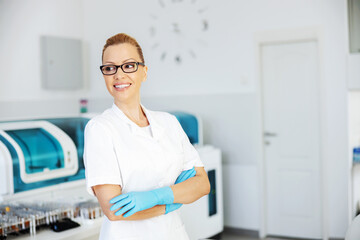 Smiling attractive positive blond female lab assistant in sterile uniform with rubber gloves on standing in laboratory with arms crossed and looking away.