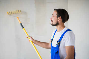 Workman in blue overalls painting the wall with a roller, view from the side