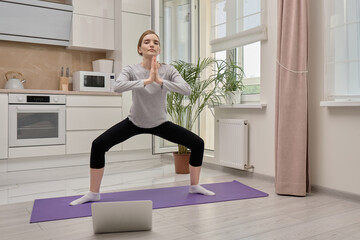 A blonde woman does yoga at home on a yoga Mat and looks at the camera. Training in sports and yoga online.