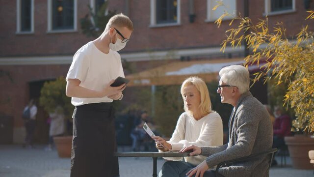Waiter in mask recording order of senior guests couple on notepad in street cafe