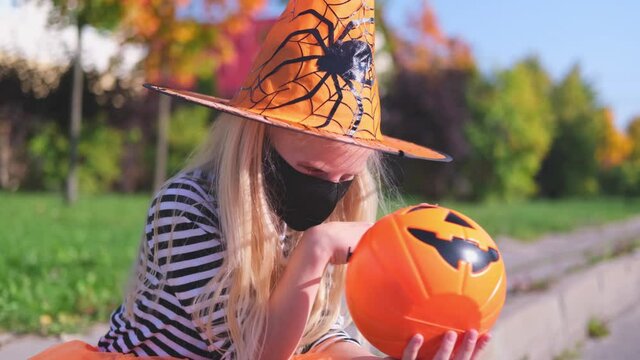 4k. Halloween kids mask. blonde girl in witch costume and pumpkin bucket. Child wearing black face mask outdoors protecting from COVID-19.