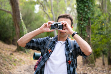 Caucasian man taking photo of nature with camera and standing on forest road. Young male traveler walking or hiking in forest. Blurred background. Tourism, adventure and summer vacation concept