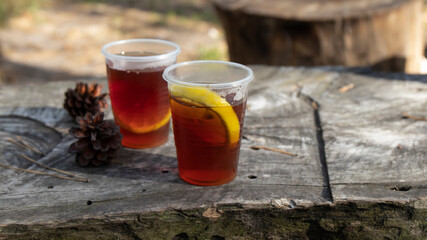 Two plastic cups of fresh hot tea with lemon stand on a weathered wooden bench in the woods. Enjoy healthy and delicious drink in the fresh air among the trees.