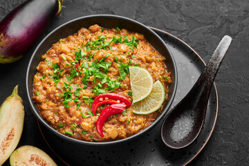 Baingan Bharta or Roasted Mashed Eggplant in black bowl on dark slate table top. Bhurta is indian cuisine puree dish of fire roasted eggplant and masala.