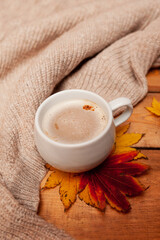 vertical cozy picture of Breakfast, coffee with milk foam in a white mug, autumn red and yellow maple leaves and a knitted beige sweater on a wooden table,selective focus