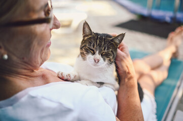 Happy smiling senior elderly woman in glasses relaxing in summer garden outdoors hugging domestic tabby cat. Retired old people and animals pets concept - Powered by Adobe