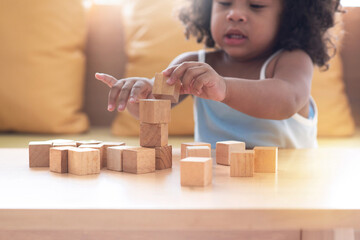 Cute dark-skinned child girl playing stacking wooden block in living room, blank wooden blocks