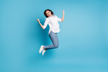 Fototapeta na wymiar Full length photo of crazy lady jumping high up rejoicing raise fists wear white shirt jeans shoes isolated blue color background