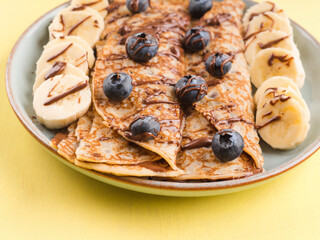 Delicious crepes filled with chocolate hazelnut spread and topped with banana and blueberries. Closeup of the dish on yellow background.