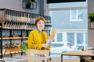 Minimalist vegan style girl with wicker bag and reusable glass coffee cup on background of interior of zero waste shop. Woman doing shopping without plastic packaging in plastic free grocery store.