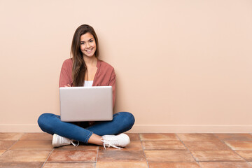 Teenager student girl sitting on the floor with a laptop keeping the arms crossed in frontal...