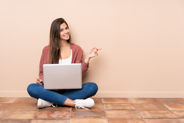 Teenager student girl sitting on the floor with a laptop pointing finger to the side