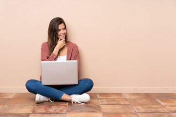 Teenager student girl sitting on the floor with a laptop looking to the side