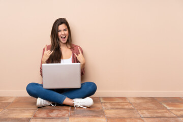 Teenager student girl sitting on the floor with a laptop making rock gesture