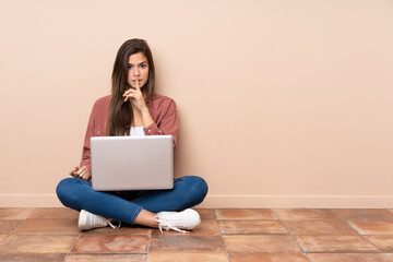Teenager student girl sitting on the floor with a laptop showing a sign of silence gesture putting finger in mouth