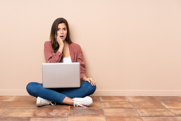 Teenager student girl sitting on the floor with a laptop surprised and shocked while looking right