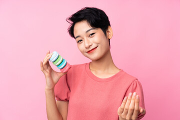 Young Asian girl over isolated pink background holding colorful French macarons and inviting to come