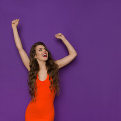 Carefree Young Woman In Orange Fitted Dress Is Holding Arms Raised And Laughing