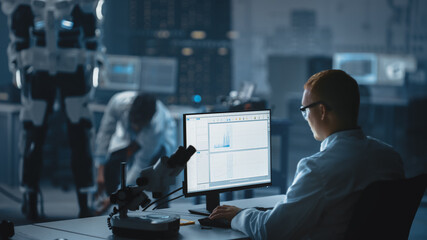 In Robotics Development Laboratory: Chief Analyst Uses Computer, Top Male Scientist Work on Bionics Exoskeleton Prototype. Designing Powered Exosuit to Help Disabled People. Over the Shoulder Shot.