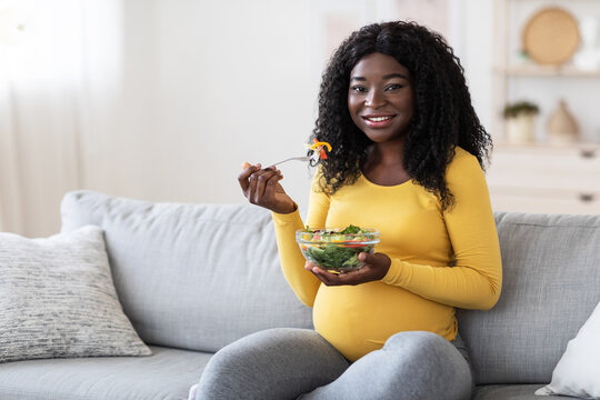 Smiling pregnant black woman eating fresh salad, sitting on sofa