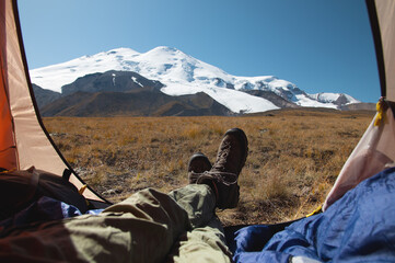 Fototapeta premium View of Mount Elbrus from an open tent with the legs of a tourist in mountain trekking boots