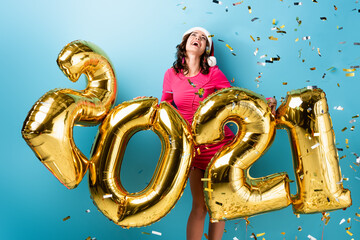 young joyful woman in santa hat near balloons with 2021 numbers and confetti on blue, new year concept