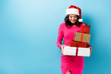 young joyful woman in santa hat and dress holding wrapped presents on blue
