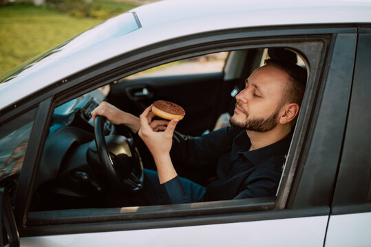 Side View Of A Young Caucasian Businessman Driving A Car On The Way To Work And Eating A Delicious Doughnut With Chocolate Cream. Sweet Breakfast In The Car