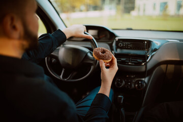 Close - up of  a caucasian businessman holding a delicious chocolate doughnut in his hand and driving a car on the way to work. Snack in the car